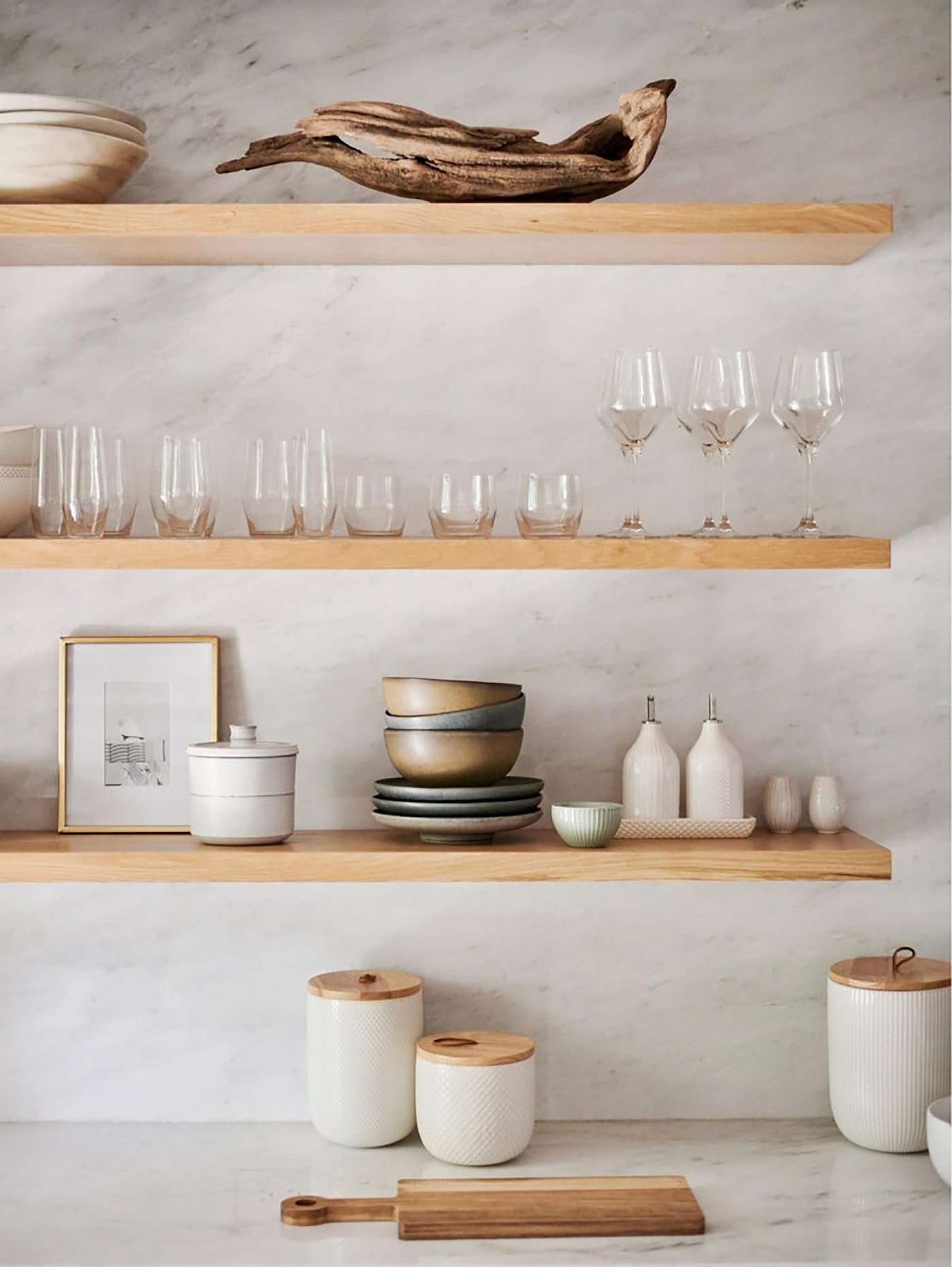 Interior still life shot of dishware, glassware, and decorative items on light wooden shelves against a textured white wall.