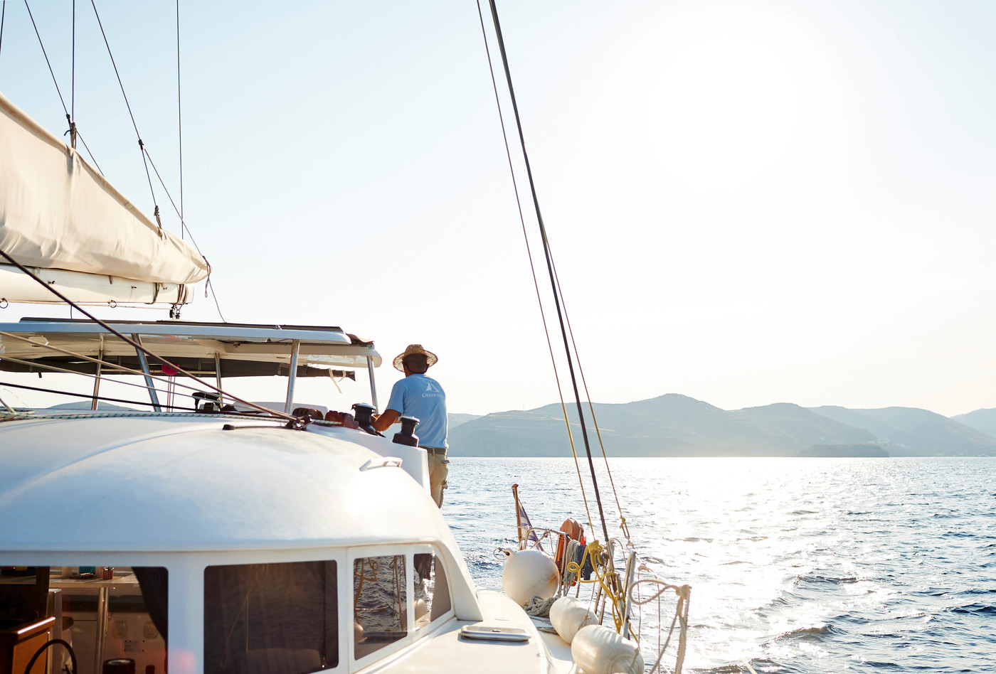 Person piloting a white sailboat on a sunny day with views of blue water and distant mountains, showcasing travel and leisure.