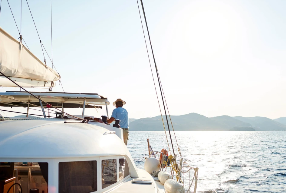 Person piloting a white sailboat on a sunny day with views of blue water and distant mountains, showcasing travel and leisure.