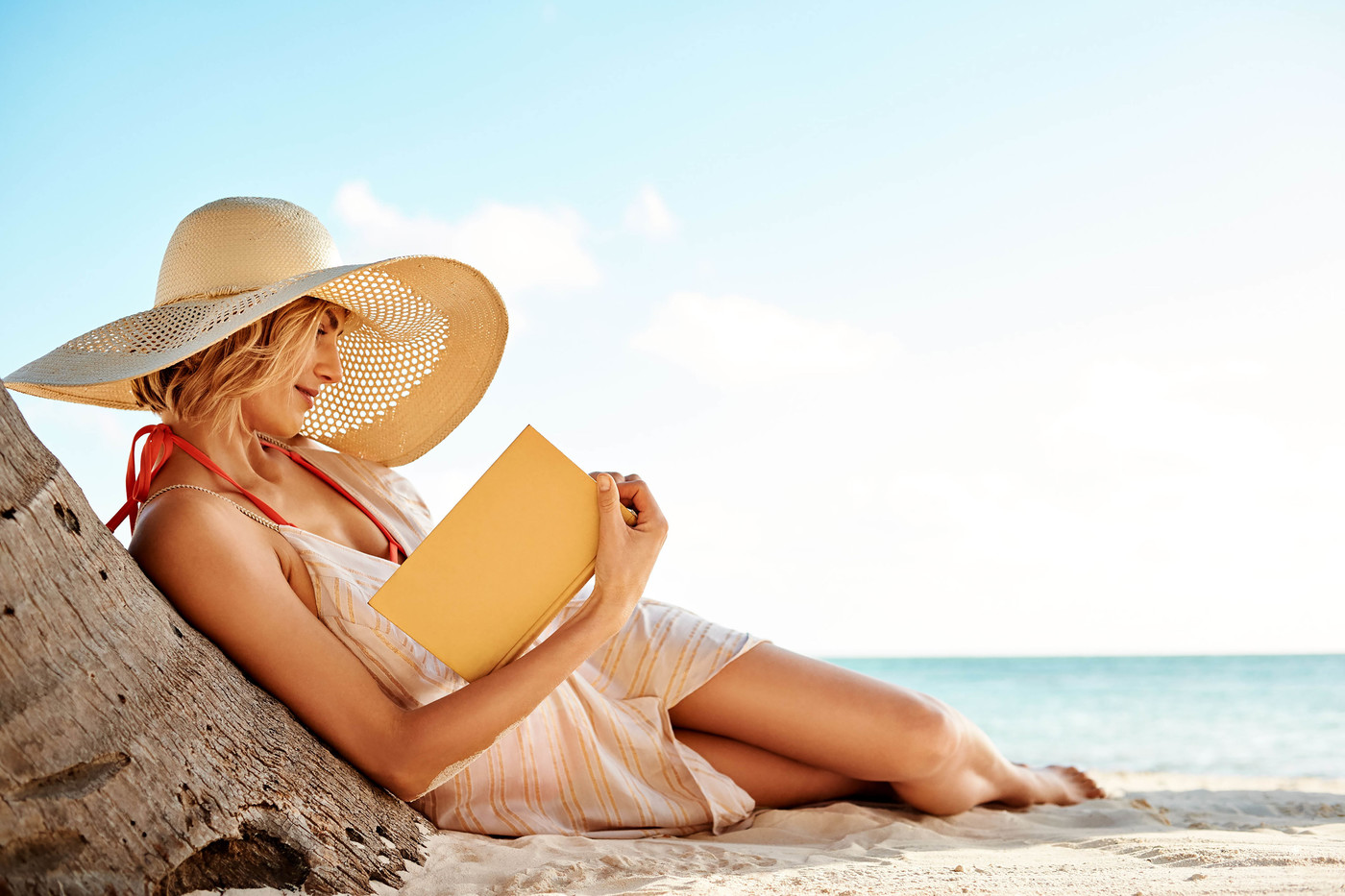 A woman in a sunhat lounges on a sandy beach next to a tree trunk, deeply engrossed in reading a book on a bright, sunny day.