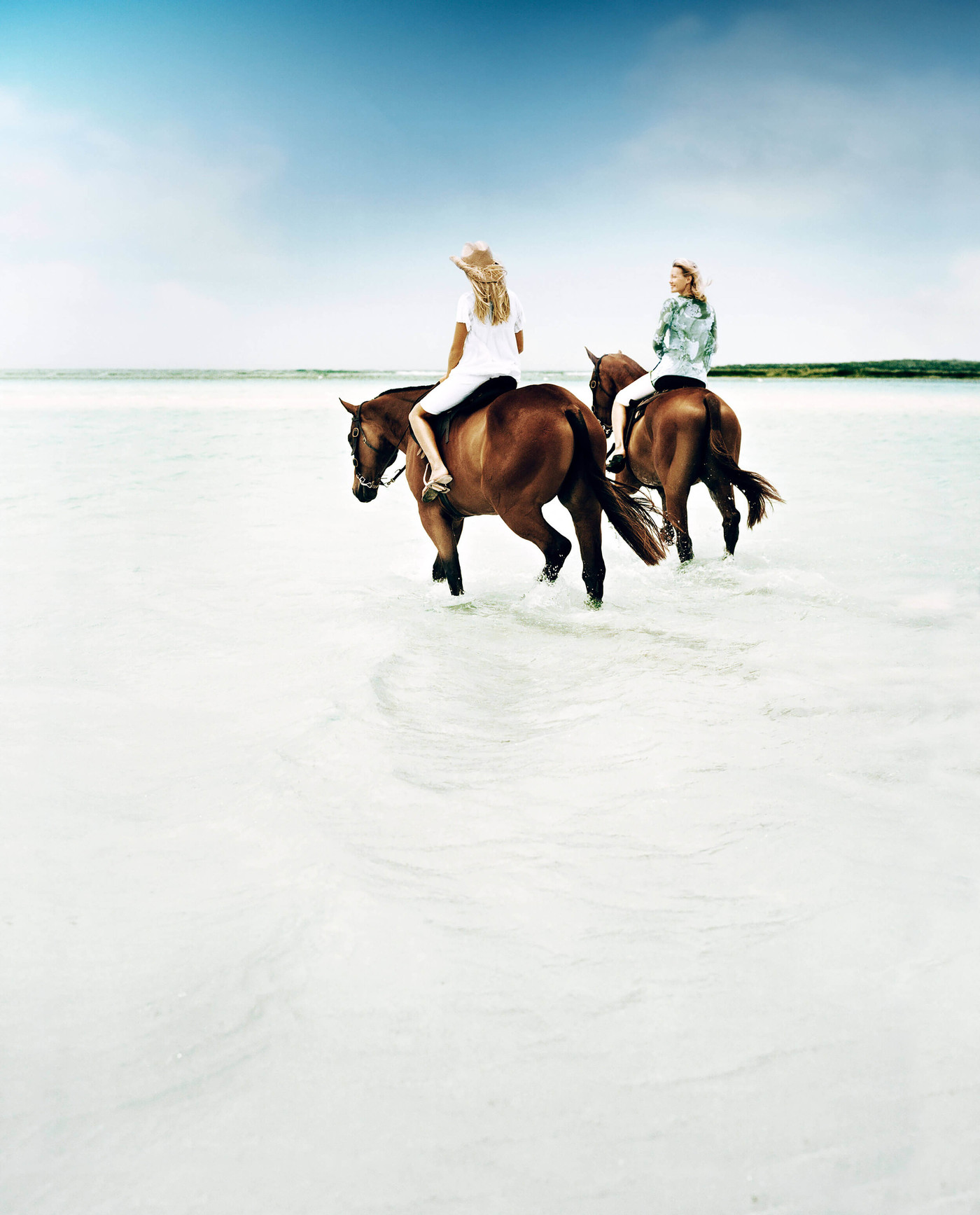 Two blonde women riding horses through shallow water at the beach on a sunny day under a blue sky with white clouds.