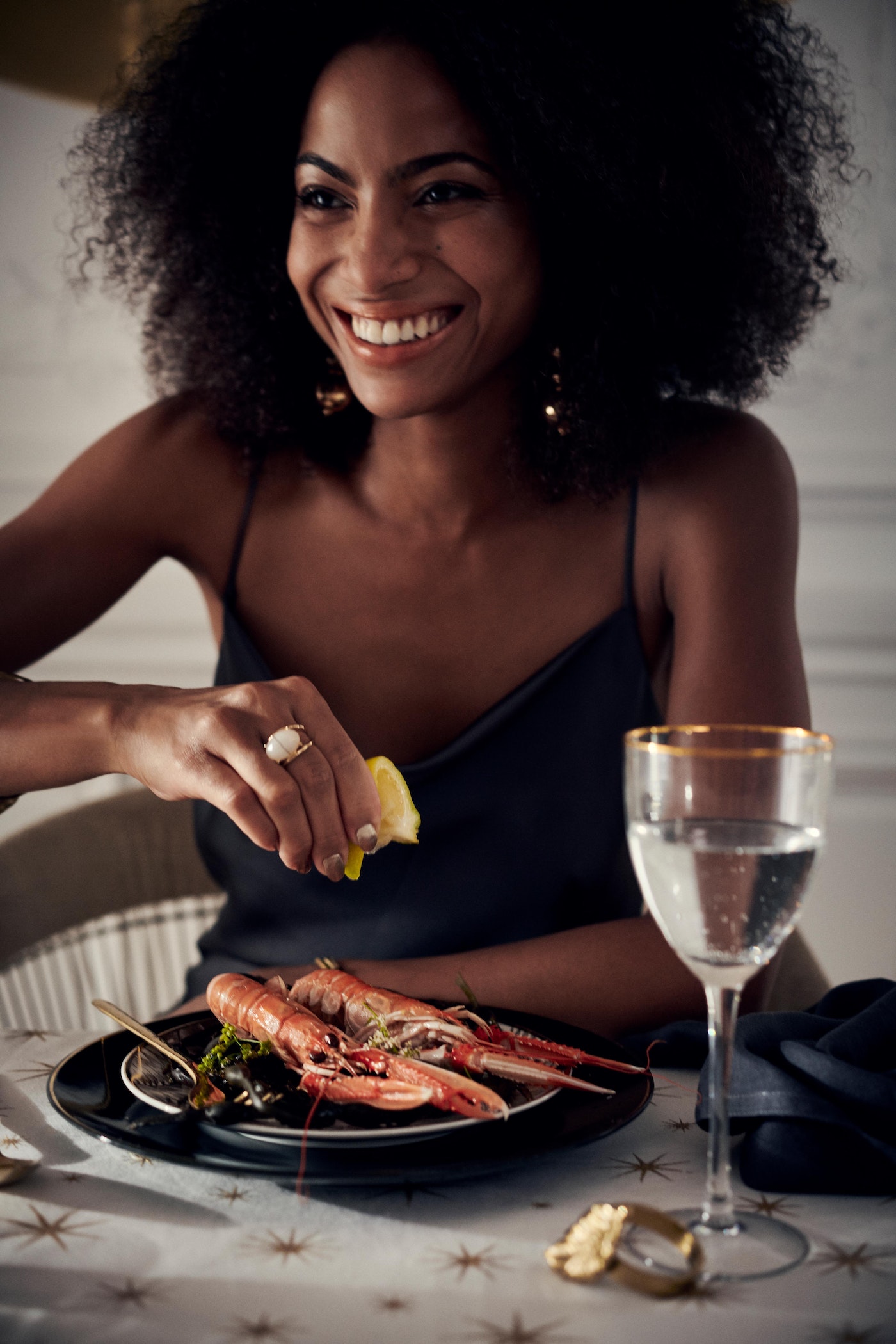 Smiling woman squeezes lemon over langoustines at table. Seafood meal, dark hair, gold jewelry, gold rimmed glass, white tablecloth.