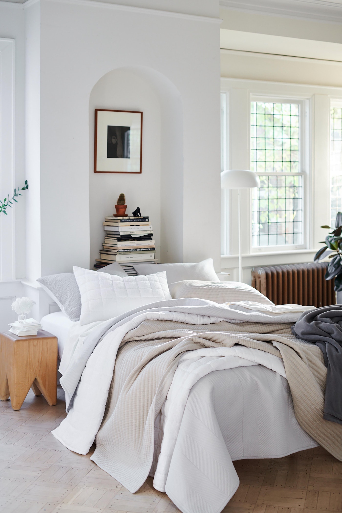 Bright and airy bedroom showcases a bed with white and beige bedding, a wooden side table, and a stack of books in a naturally lit interior.