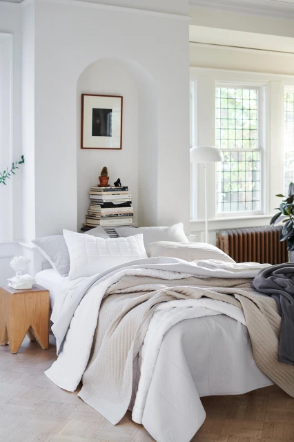 Bright and airy bedroom showcases a bed with white and beige bedding, a wooden side table, and a stack of books in a naturally lit interior.