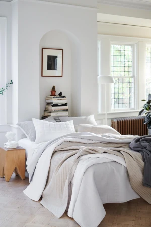 Bright and airy bedroom showcases a bed with white and beige bedding, a wooden side table, and a stack of books in a naturally lit interior.