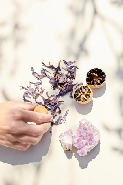 Overhead shot of a hand arranging dried violet flowers, amethyst crystals, and dehydrated citrus fruit on a light marble surface.