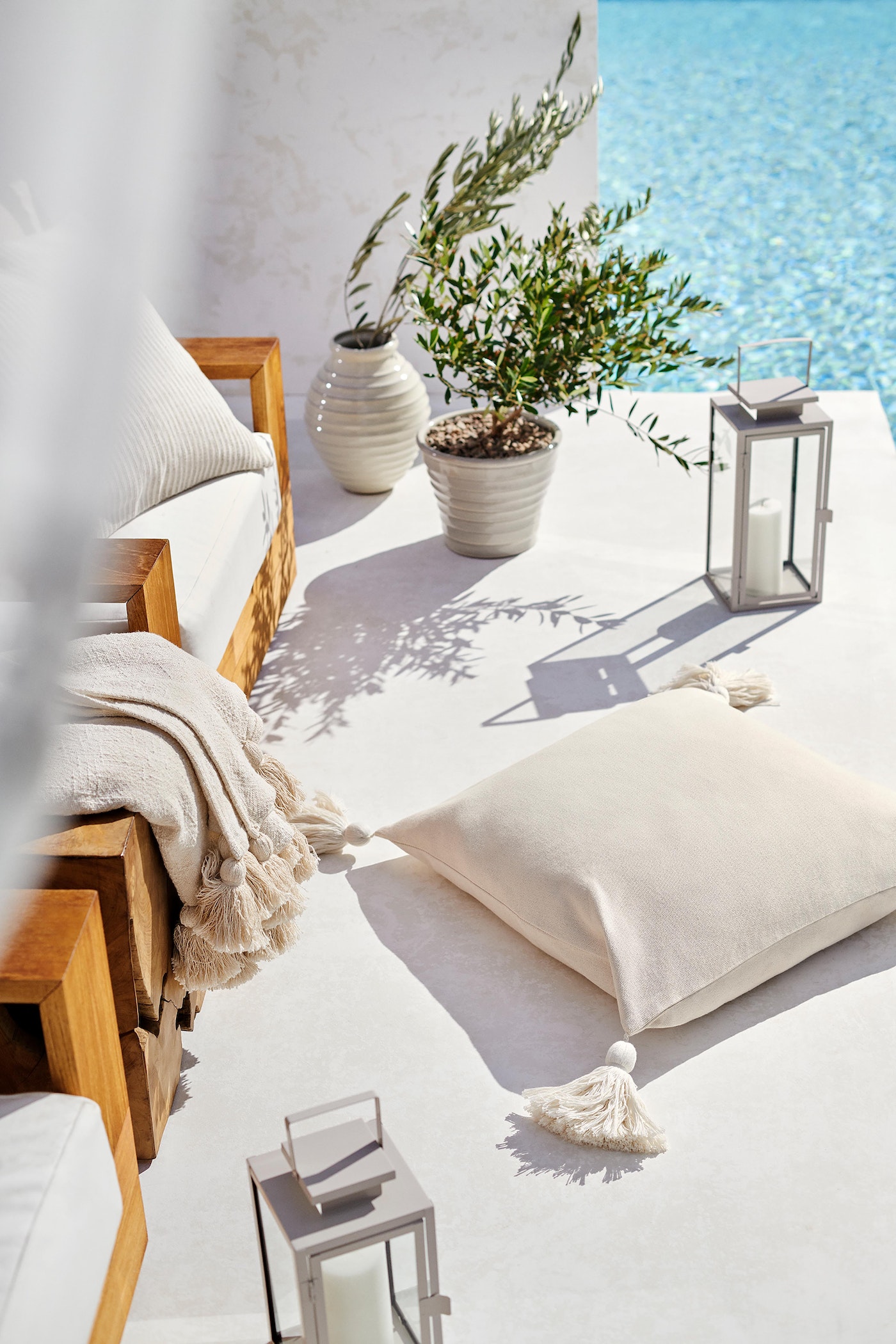 Outdoor seating area with a wooden frame sofa and off-white cushions and throw pillows beside a pool on a sunny day.