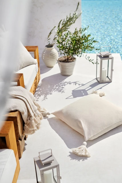 Outdoor seating area with a wooden frame sofa and off-white cushions and throw pillows beside a pool on a sunny day.
