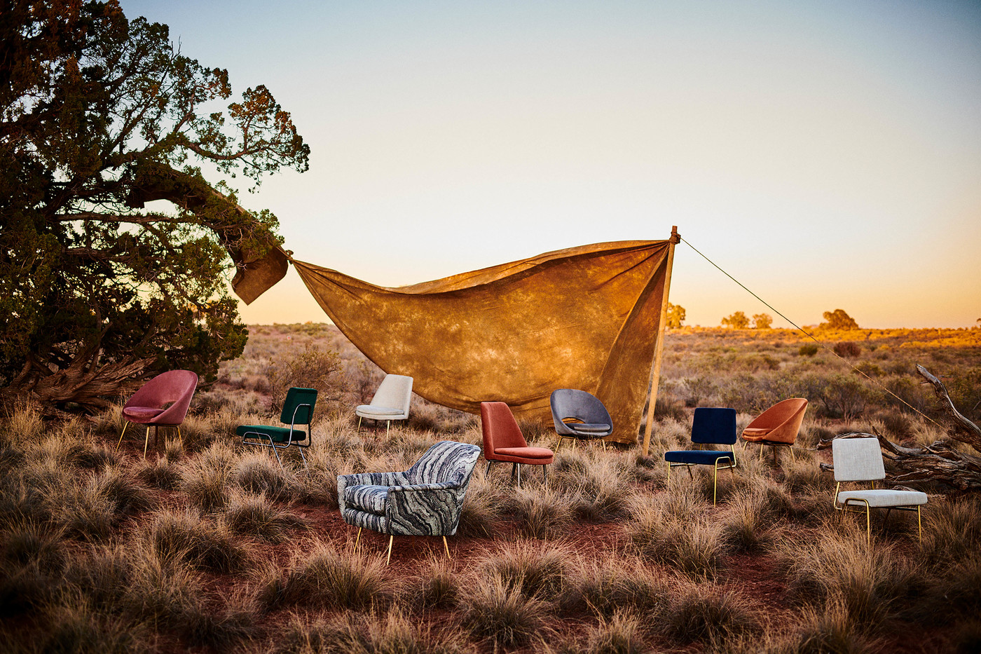 Outdoor furniture arrangement with armchairs under a draped tarp in a grassy, rural setting at sunset with warm, diffused lighting.