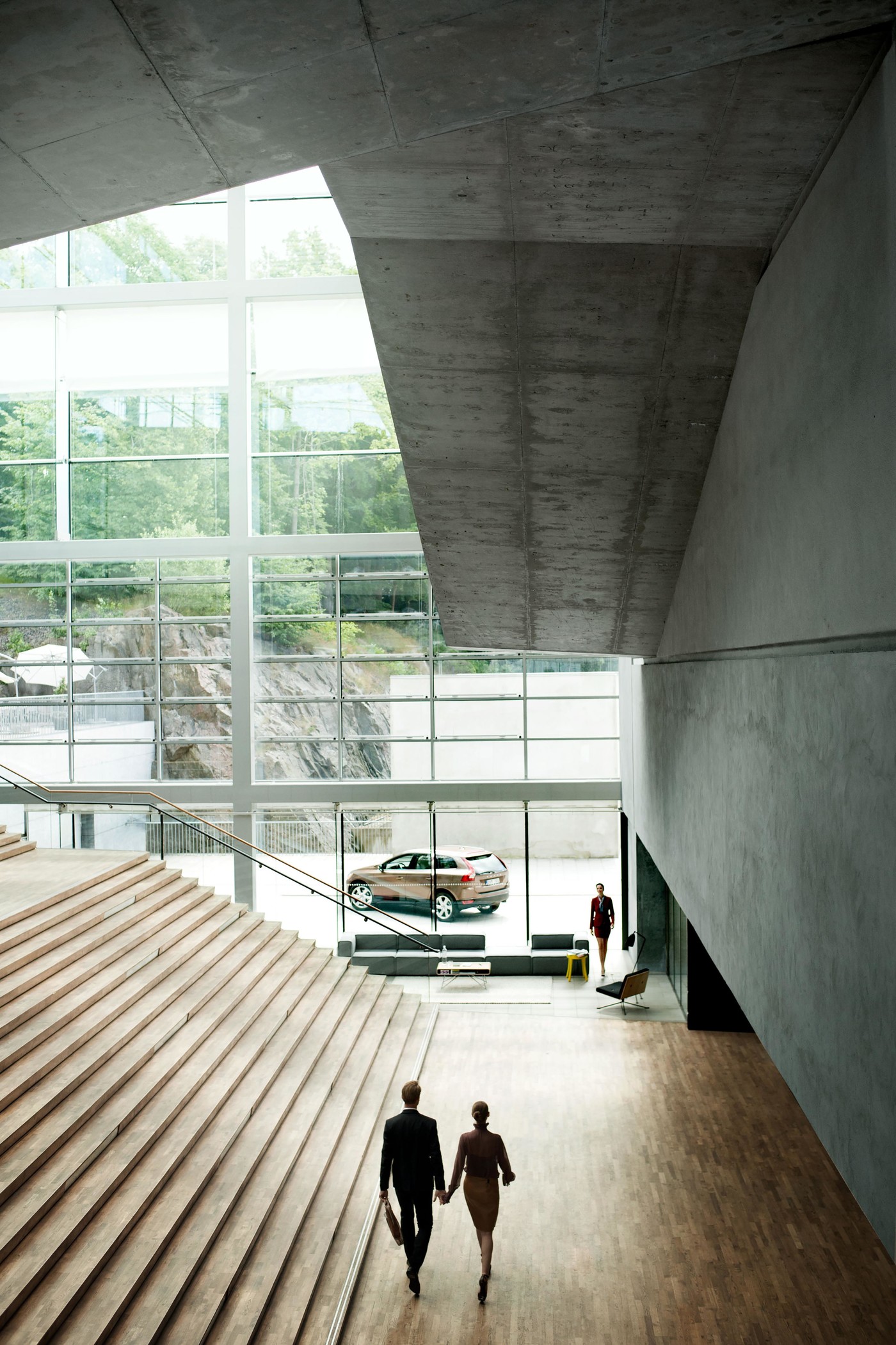 Couple in suits walking through modern building interior with car display near large windows and staircase, concrete architecture.
