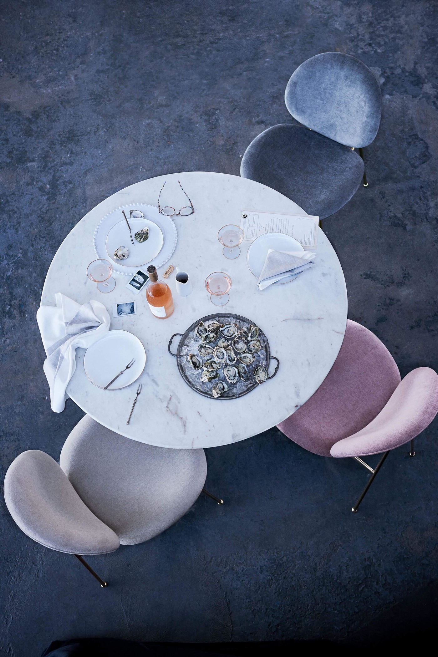 Overhead shot of a round marble table set with oysters, plates, glasses, a bottle, and silverware, surrounded by three modern chairs.