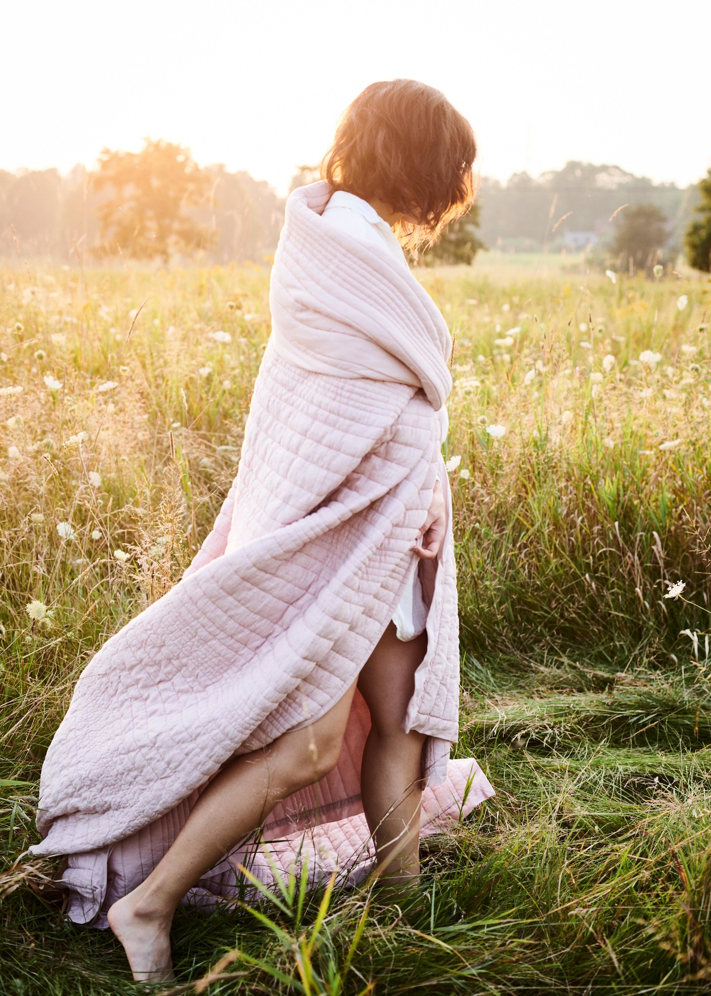 A woman is wrapped in a quilted, light pink blanket, standing barefoot in a grassy field. Sunlight softly illuminates the scene.