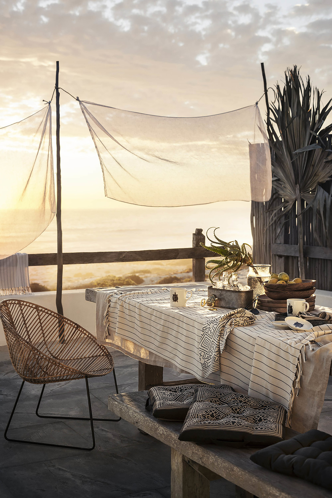 Outdoor dining area with a set table, chair, pillows, and distant ocean view under a sheer canopy at sunset with warm light.