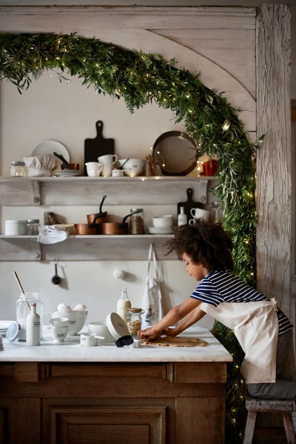 Young boy in a white apron is at a counter, cutting cookie dough, in an indoor kitchen with shelves, bowls, and holiday decorations.