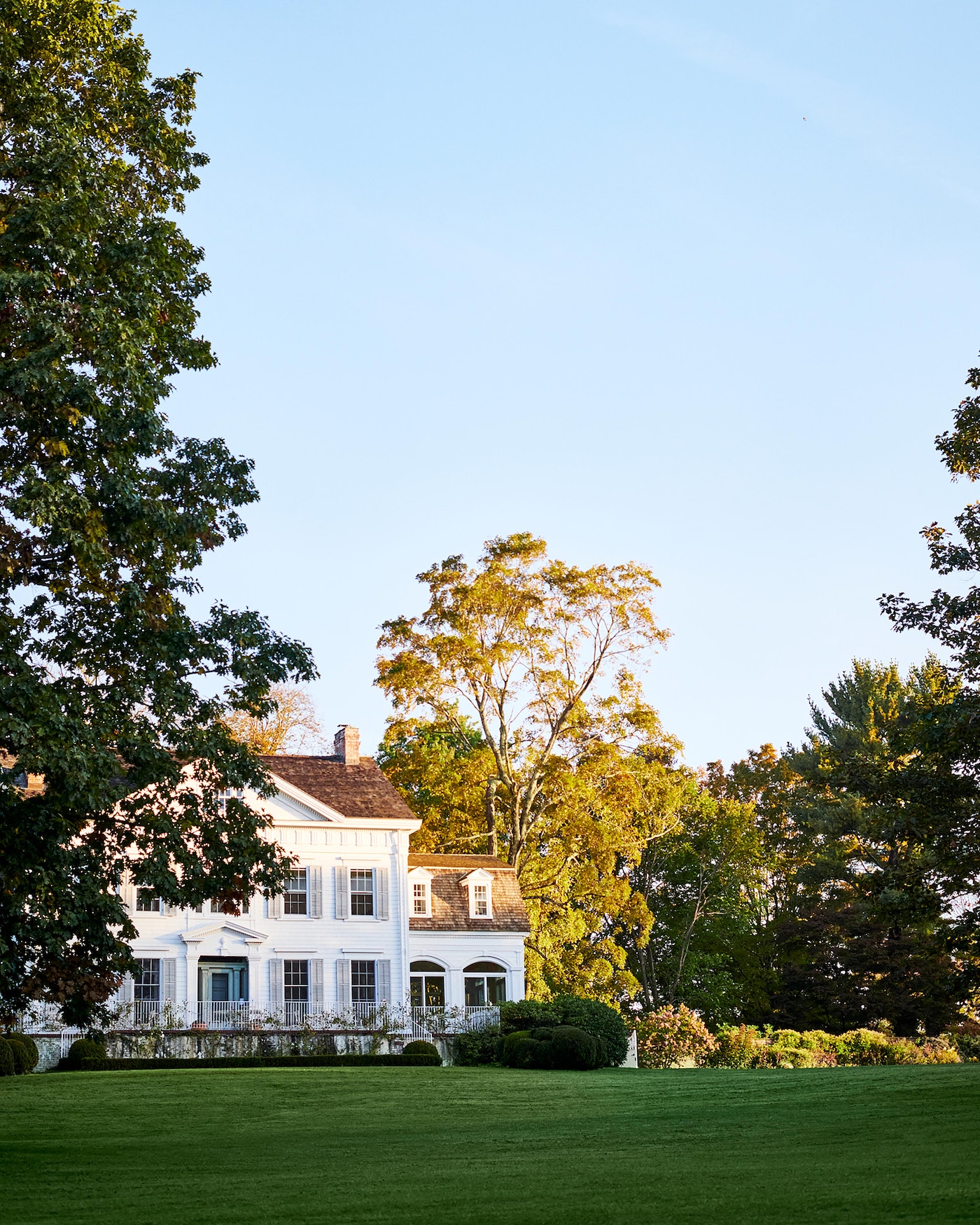 Exterior daytime view of a white, multi-story colonial-style house with green lawn and trees against a blue sky in a rural setting.