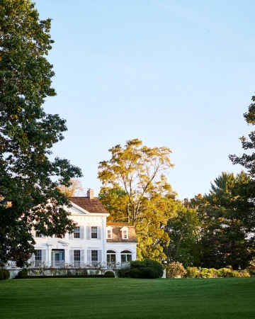 Exterior daytime view of a white, multi-story colonial-style house with green lawn and trees against a blue sky in a rural setting.