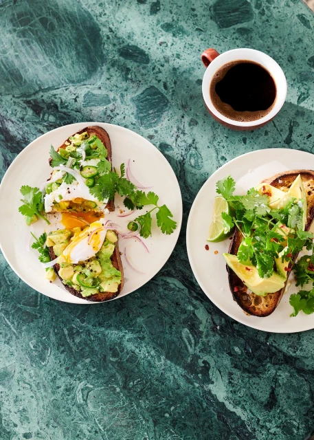 Overhead shot of two plates of avocado toast with eggs, cilantro, and lime, plus coffee cup on green marble table.