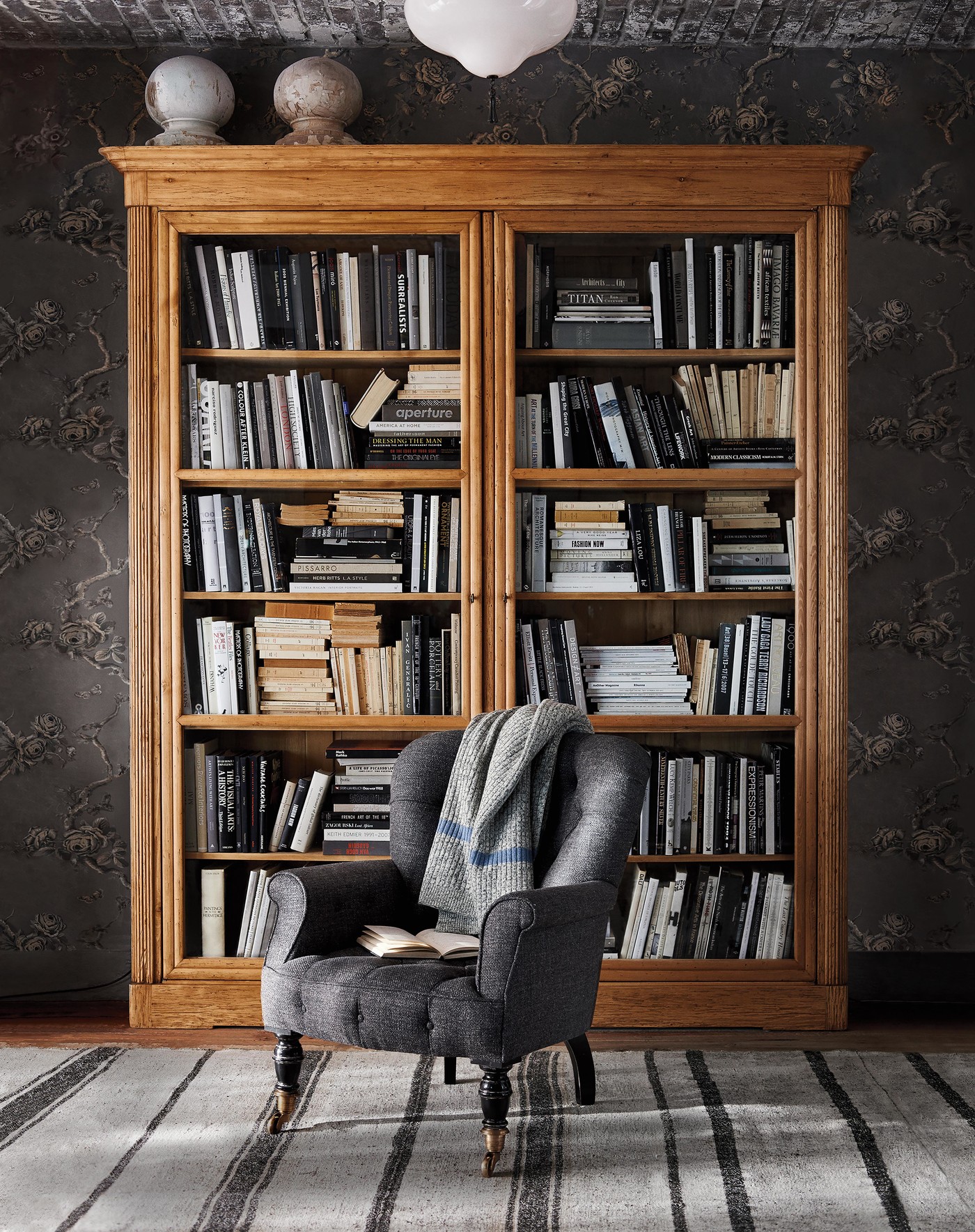Grey tufted armchair with a blanket and book sits in front of a wooden bookcase filled with books, resting on a striped carpet indoors.