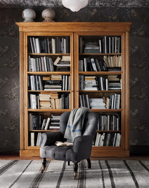 Grey tufted armchair with a blanket and book sits in front of a wooden bookcase filled with books, resting on a striped carpet indoors.