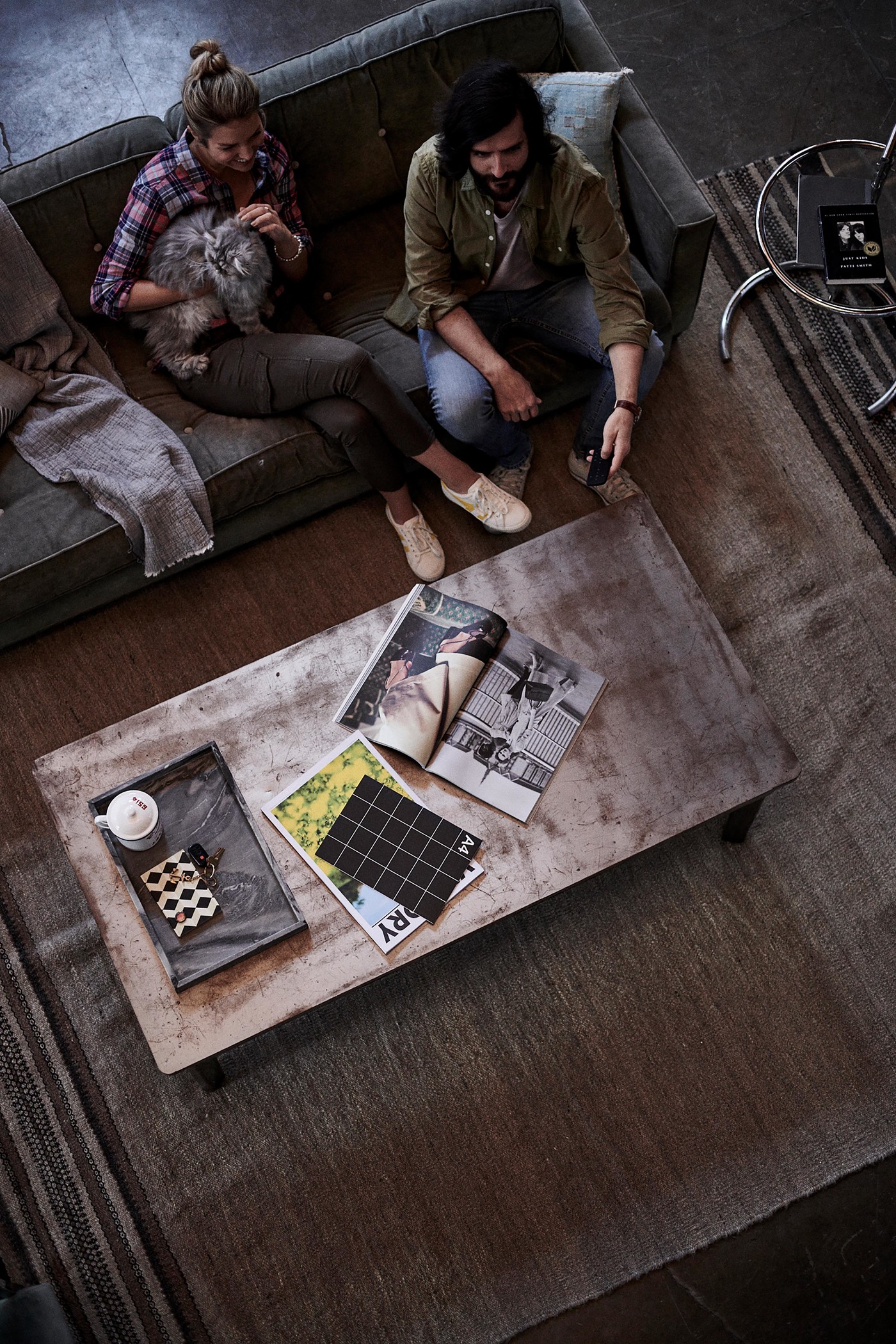 Overhead shot shows a man and woman on a couch with a cat, a coffee table holding magazines, and a patterned rug on the floor.