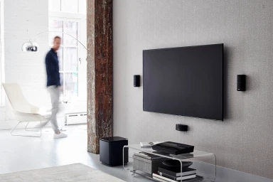 Modern living room interior with a large wall-mounted flat screen television, surround speakers, and an acrylic coffee table.