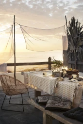 Outdoor dining table set with patterned linens under sheer fabric shades overlooking the ocean at sunset.