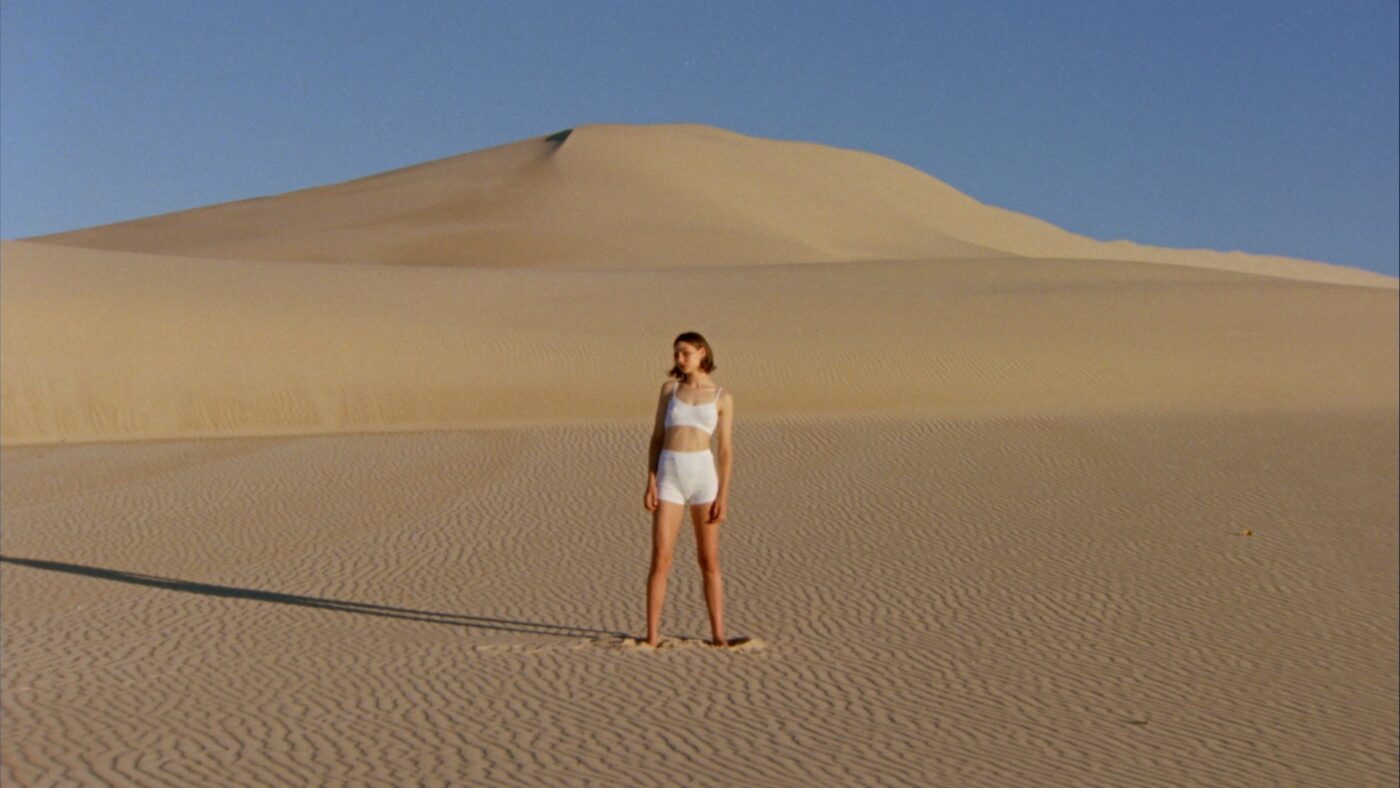 Woman in white apparel stands on sand dune against sand dune landscape and clear sky in outdoor shot, bathed in warm, natural light.