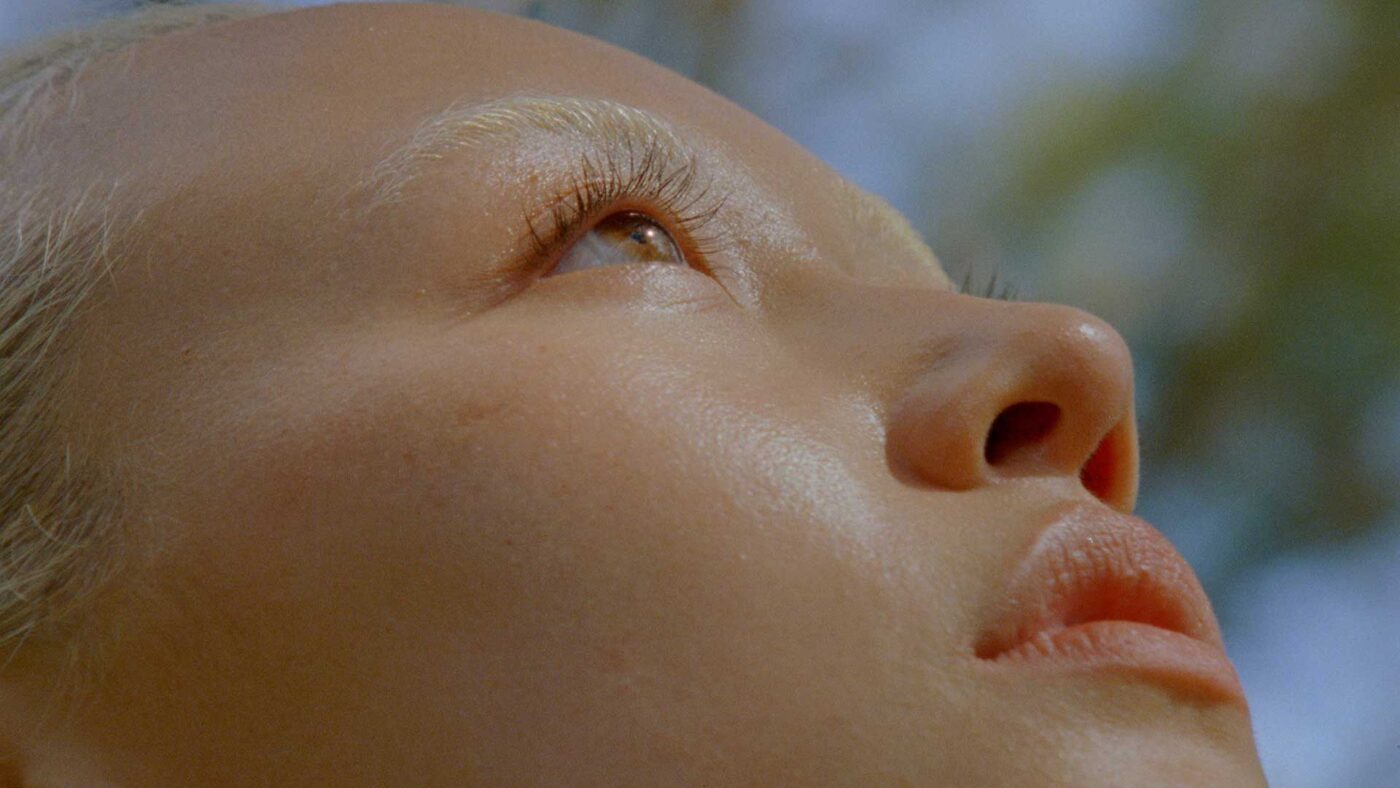 Close-up of a woman's face, focusing on her eye, nose, and lips, shot from a low angle. The blurred background suggests an outdoor setting.