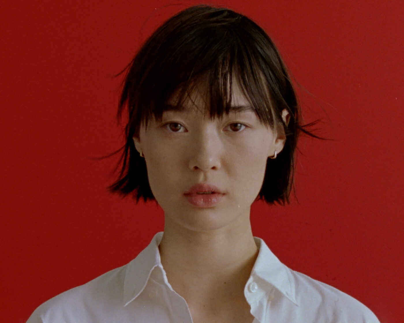 Close-up portrait of an Asian woman with short dark hair and a white collared shirt against a vibrant red backdrop.