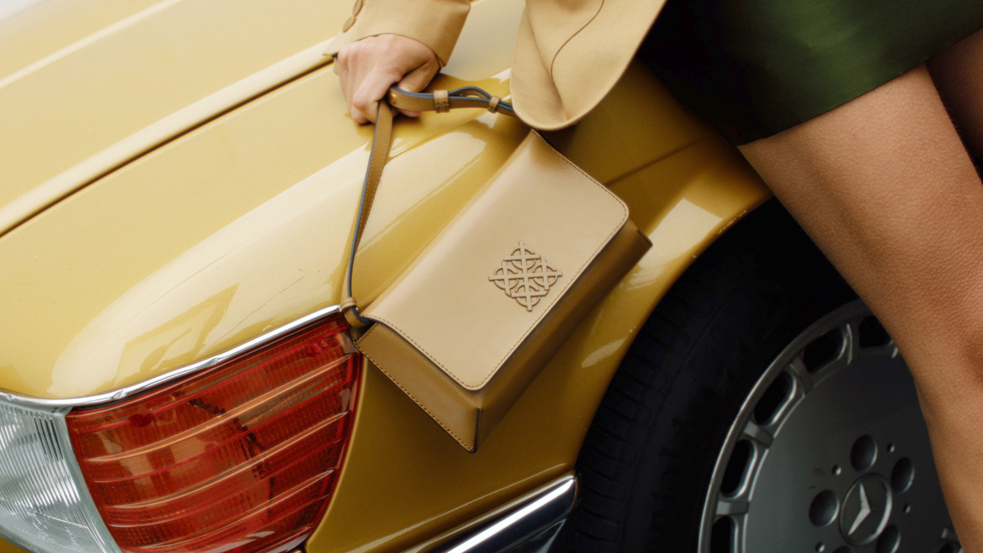 Tan-colored handbag with decorative logo displayed on the golden yellow trunk of a classic car with the wheel and rear light visible.