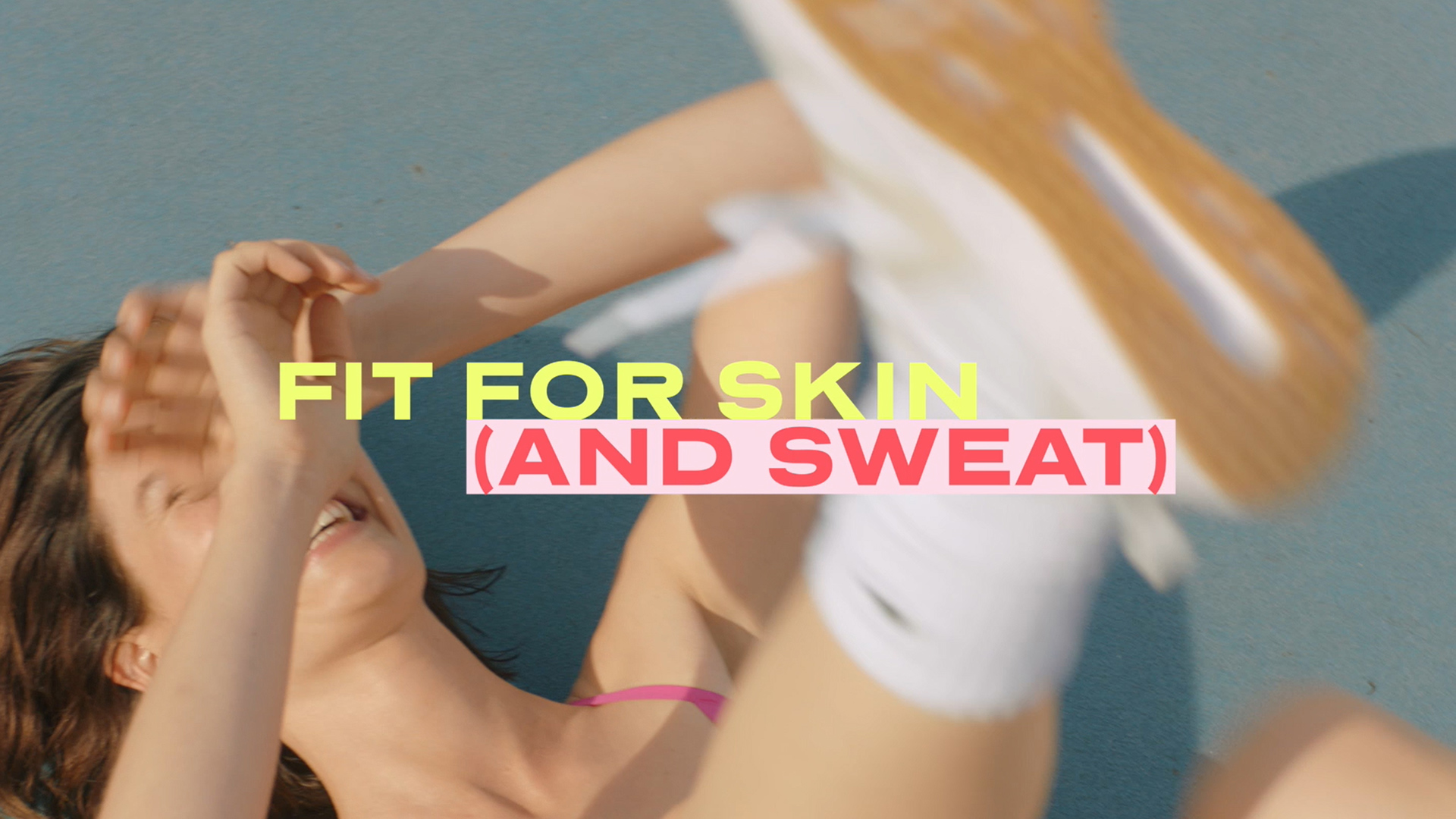 Smiling woman lying on blue surface with her arms raised, wearing athletic wear and white sneakers, with the text "FIT FOR SKIN (AND SWEAT)".