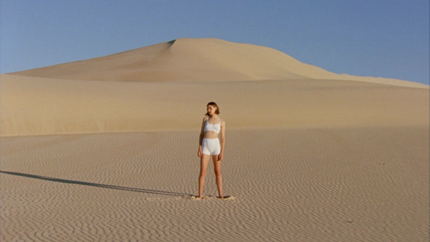 Woman in white underwear standing alone on a vast desert sand dune under a clear blue sky