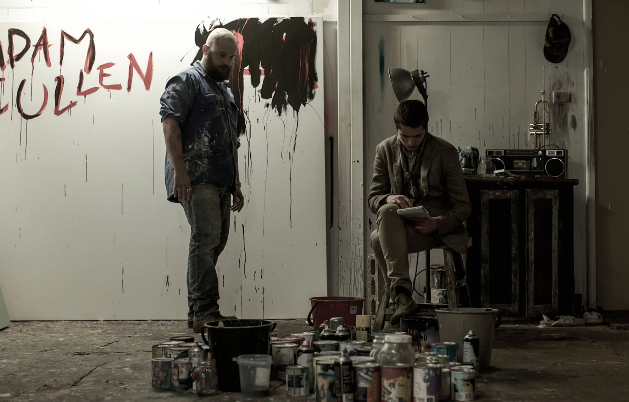 Two men in an art studio with paint cans and a large canvas with painted text, one standing and the other sitting and writing.