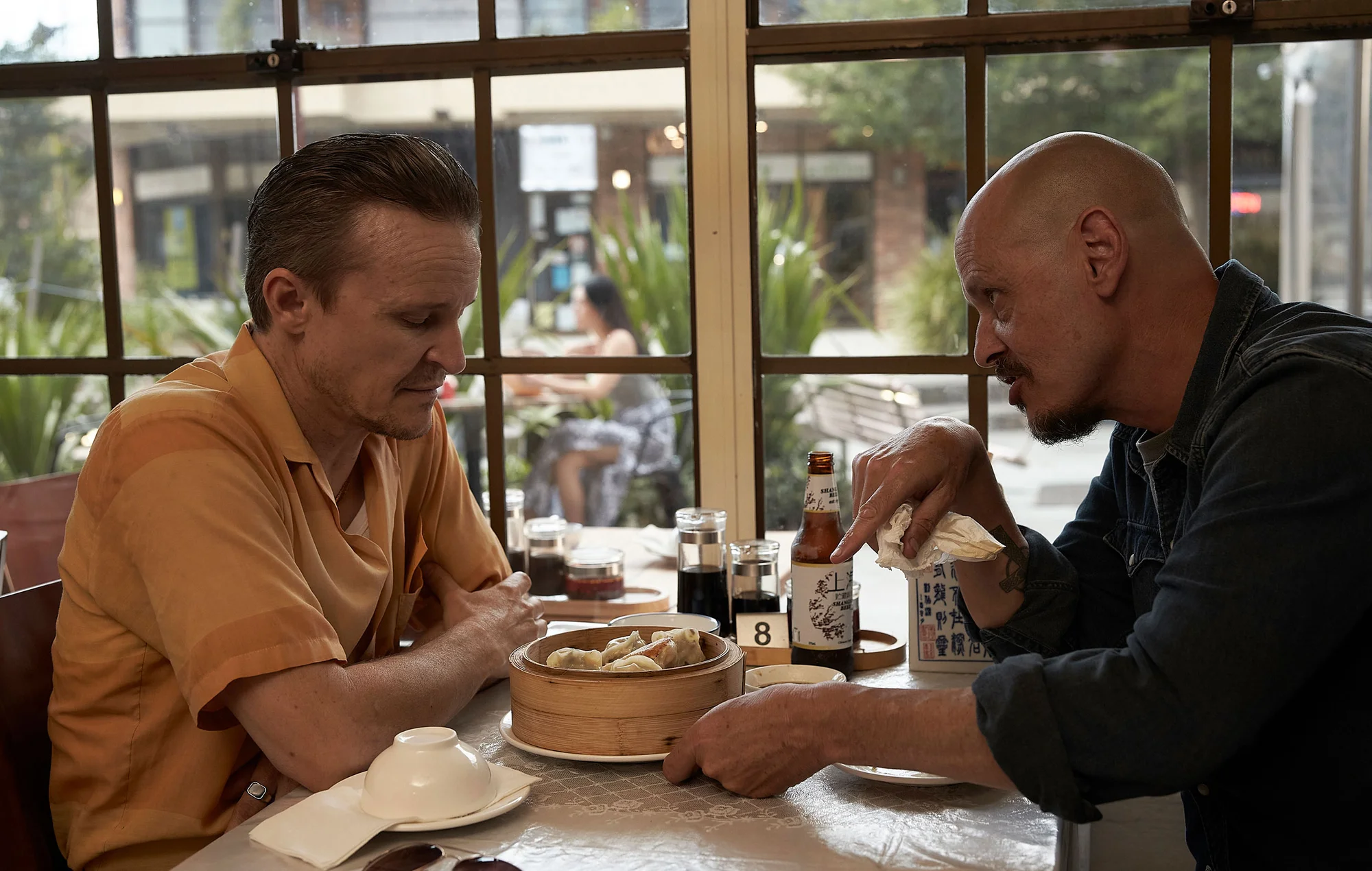Two adult men sit at a table in a restaurant, sharing a dim sum steamer, beer, and condiments with sunlight streaming in through a window.