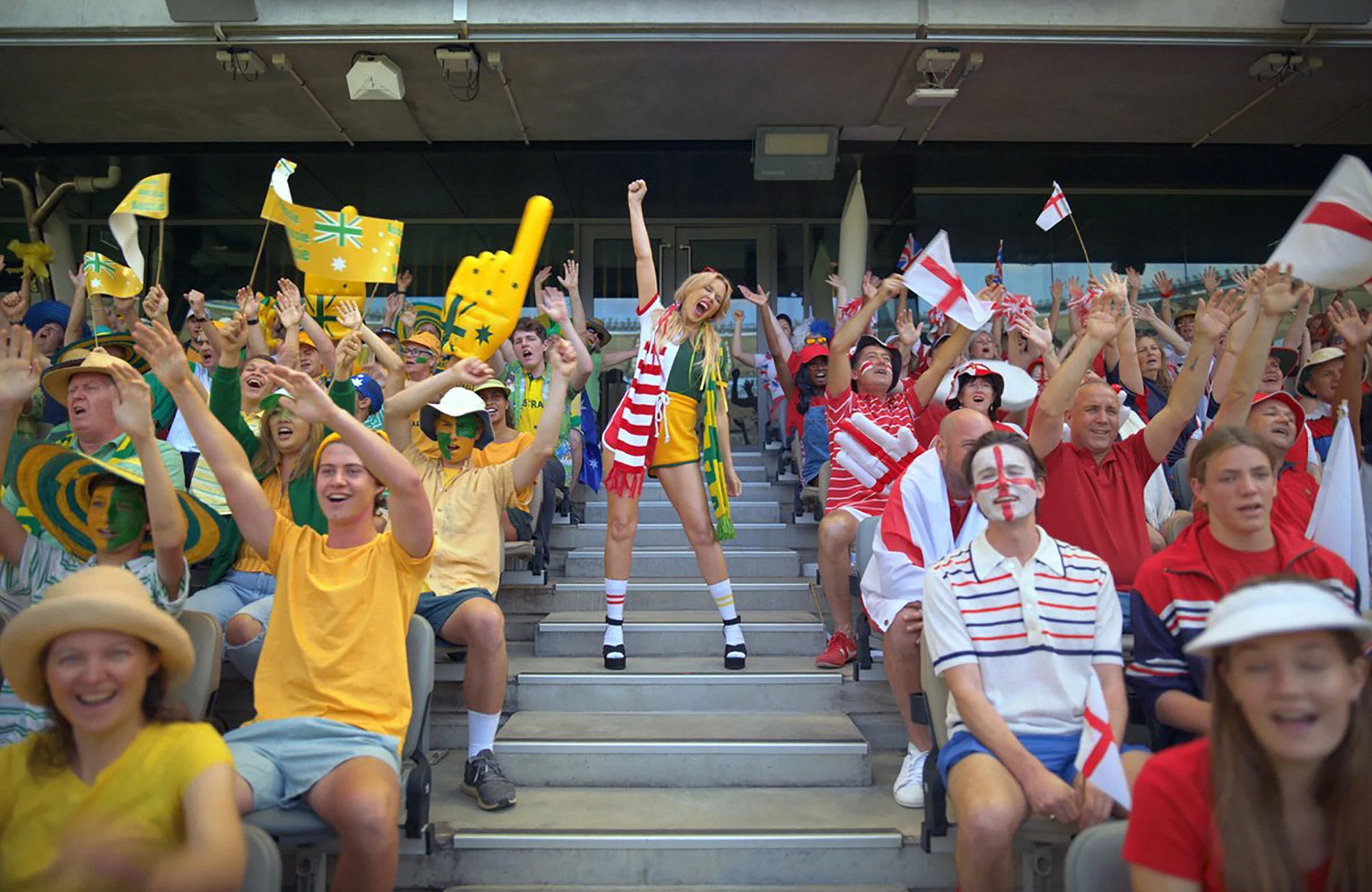 Enthusiastic sports fans cheering, waving flags and raising arms in stadium seating with a blonde woman standing in the foreground.