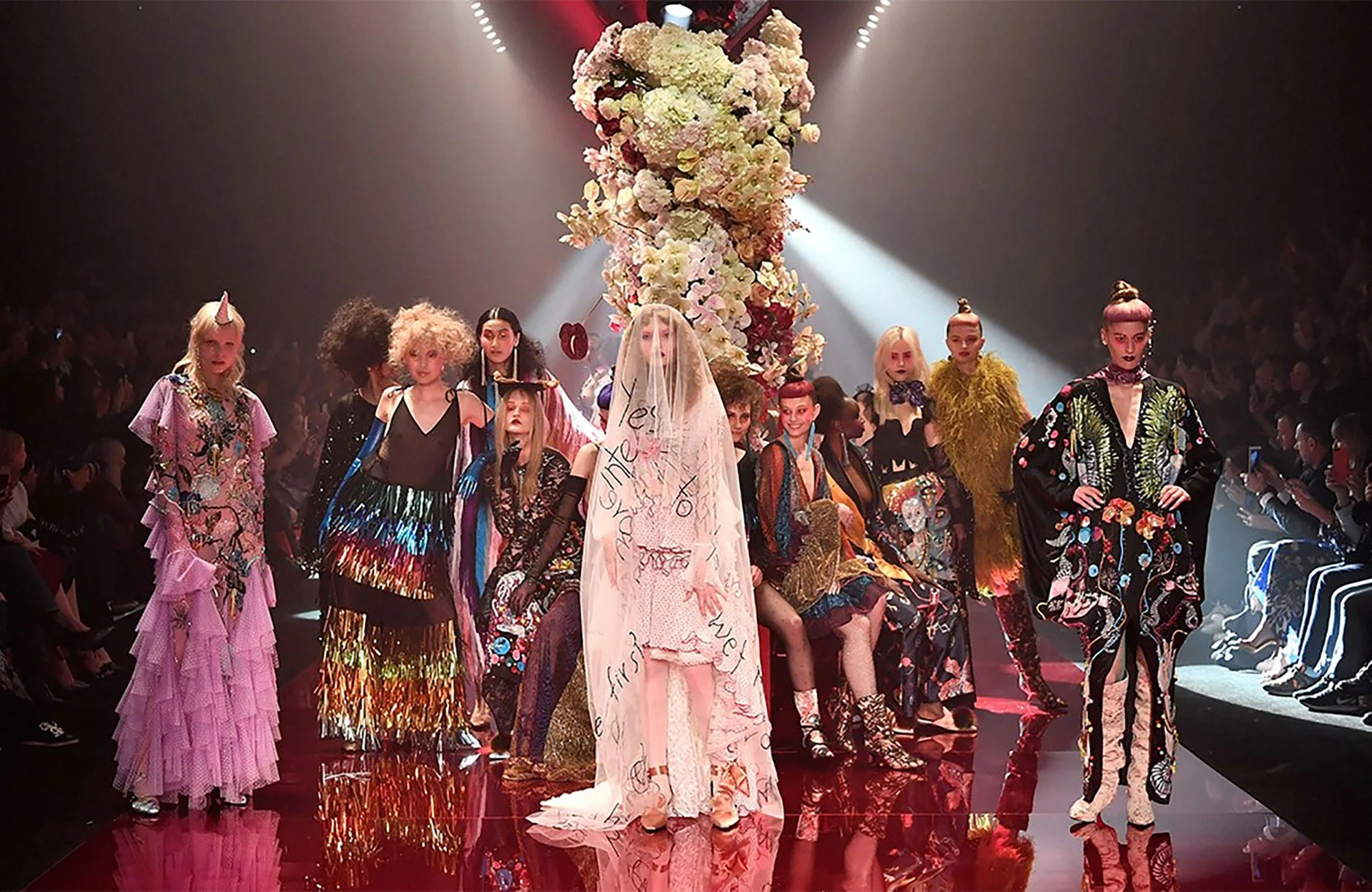 Runway fashion show features models in various colorful outfits, standing on a red reflective floor under bright spotlights and floral decor.