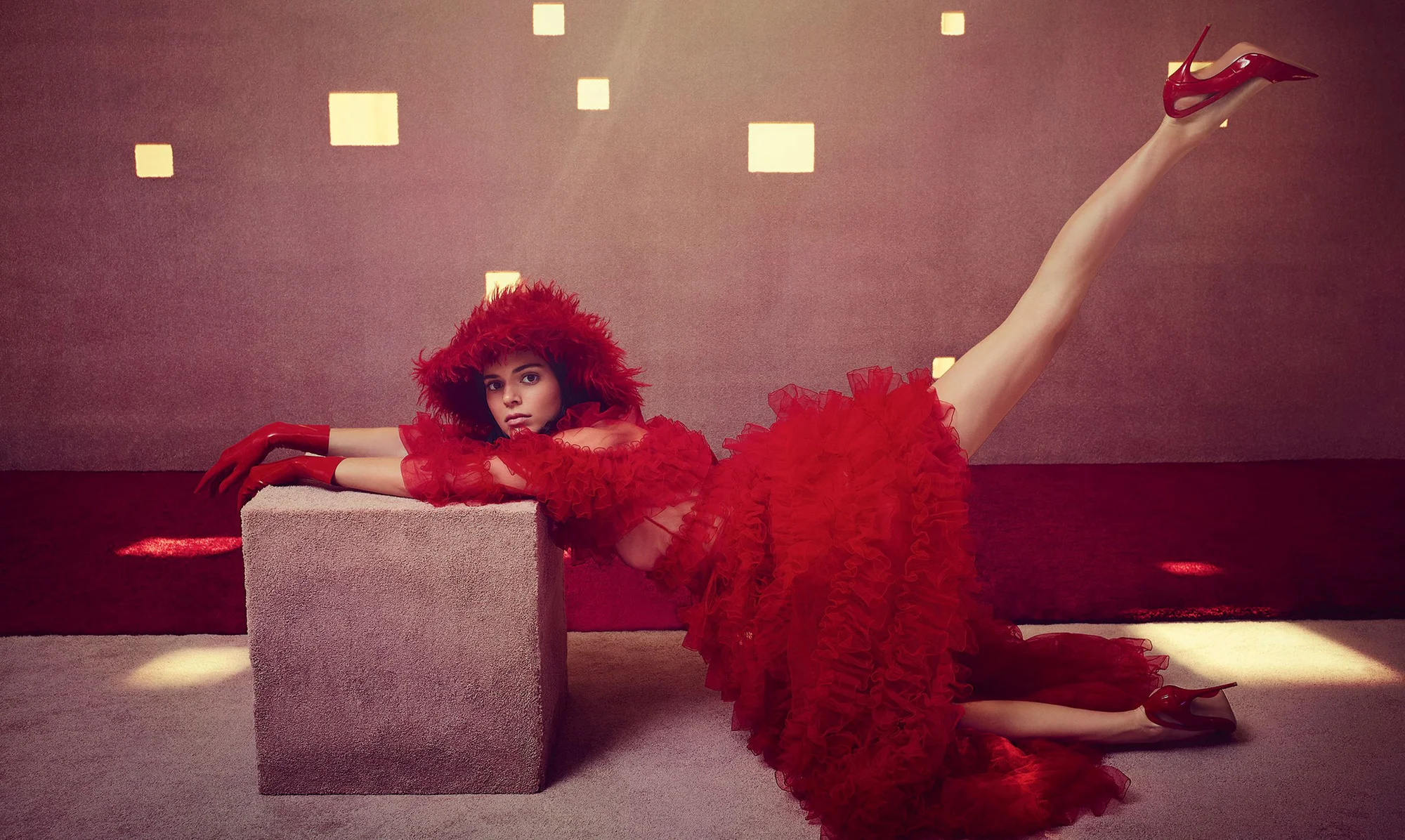 A woman in a striking red dress and matching high heels poses dramatically with one leg extended straight up, resting her arms on a cube.