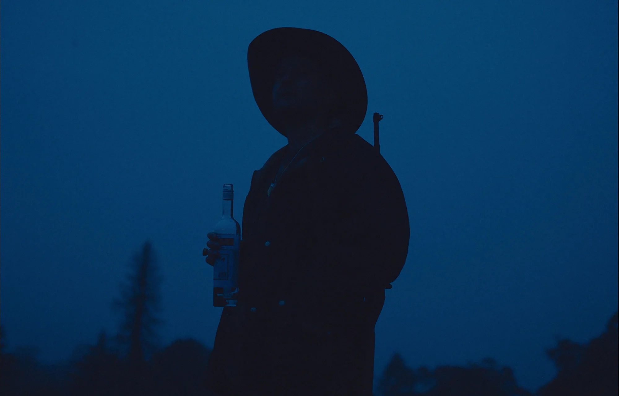 Silhouette of a person with a hat and rifle holding a bottle against a dark blue background at dusk, highlighting a moody outdoor scene.