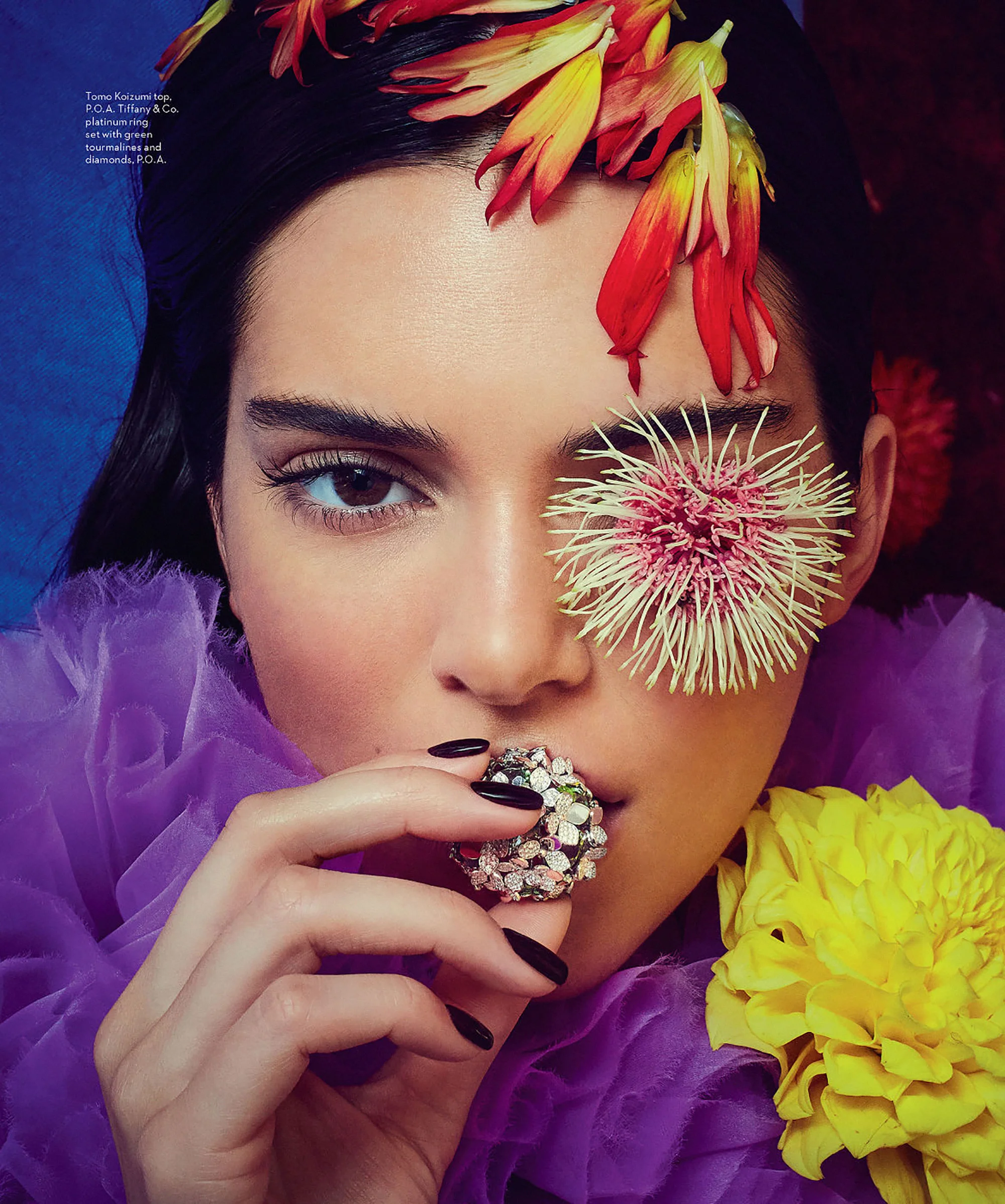 Close-up of a woman with dark hair and flowers, wearing a purple dress and holding a floral ring in front of her mouth.