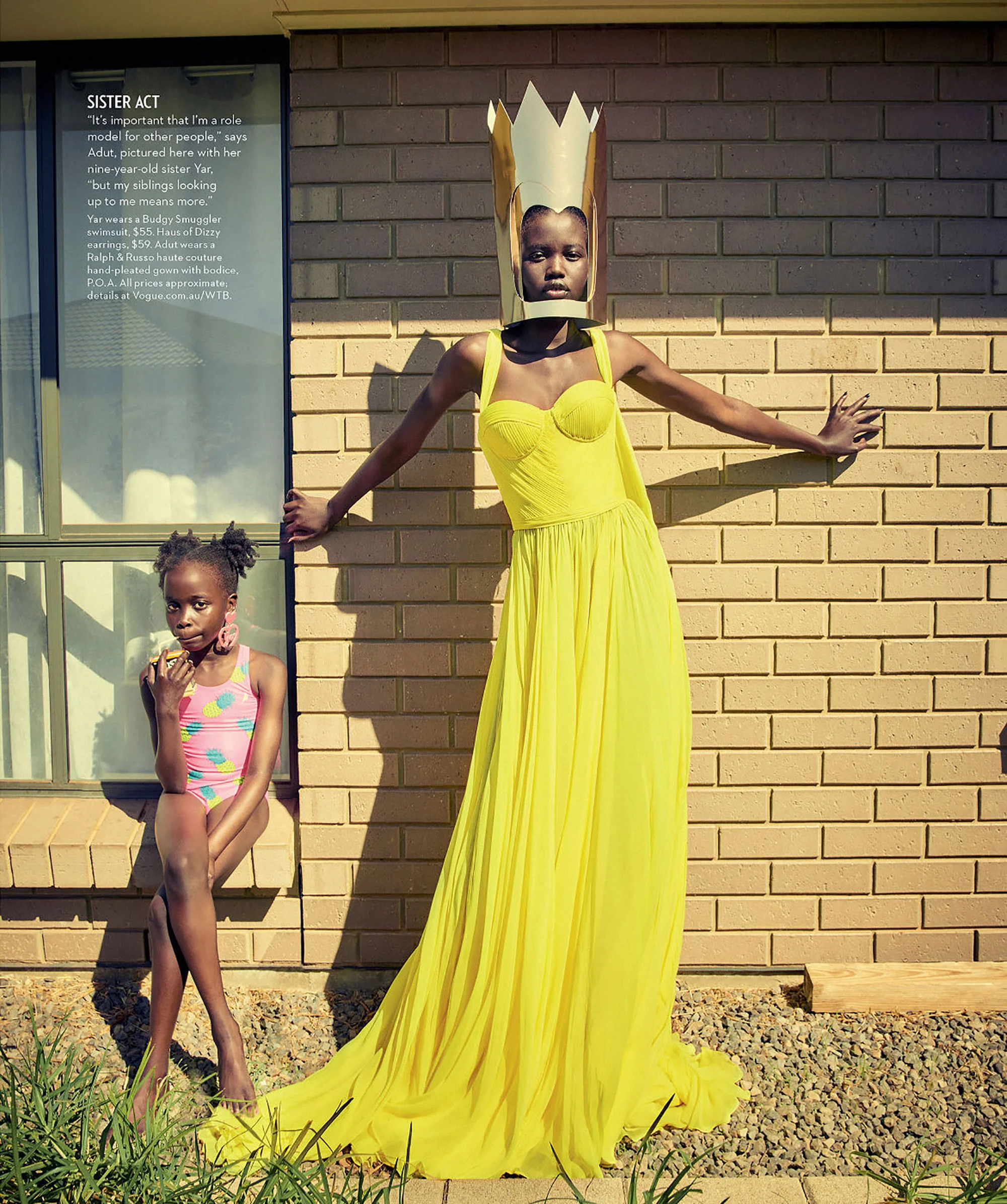 Two black girls in outdoor portrait. One stands wearing yellow dress and crown. One seated wearing swimsuit by building wall.