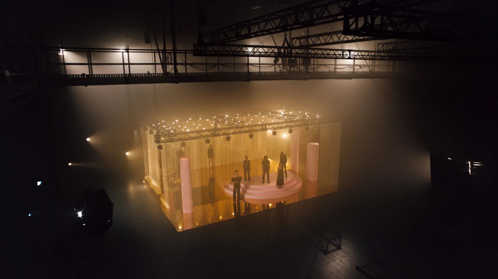 Low-angle shot of a performance stage with six people standing on it, bathed in soft light, surrounded by pink columns and golden curtains.
