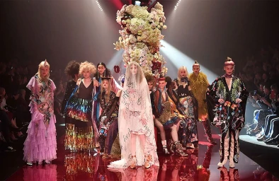 Models on a glossy runway wearing avant-garde couture and elaborate garments beneath a large hanging floral installation.