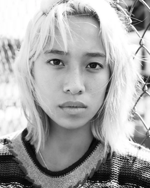 Close-up of an Asian young adult female with light-colored hair looking directly at the camera, wearing a patterned top and silver chain.