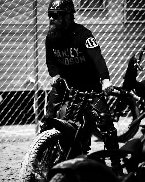 Black and white shot of a man with a long beard wearing a helmet, goggles, and Harley Davidson shirt, riding a vintage motorcycle.