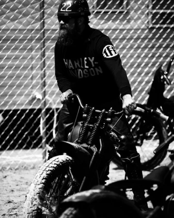Black and white shot of a man with a long beard wearing a helmet, goggles, and Harley Davidson shirt, riding a vintage motorcycle.