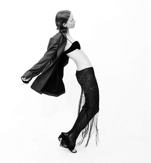 Black-and-white fashion shot of a woman in an open blazer, bra, and embellished fringe pants, arching her back against a white backdrop.