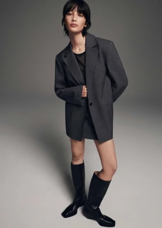 Woman wearing an oversized charcoal blazer and black knee-high boots against a plain grey studio background.