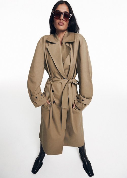 A woman in a tan trench coat and black boots poses against a white background, hands in her pockets, wearing sunglasses in a studio shot.