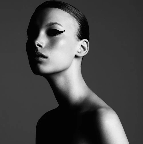 Striking black and white studio portrait of an adult woman with strong winged eyeliner, dramatic lighting, and a grey background.
