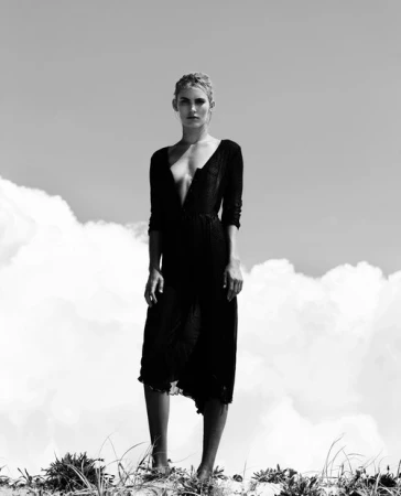 Monochrome full-length portrait of a woman in a dark V-neck dress standing against an overcast sky and grassy dunes.
